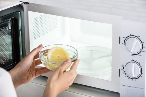 A person places a glass bowl with lemon slices and water into a microwave, preparing to deodorise it naturally. This method is often recommended by appliance experts like Quality Appliance Repairs (QAR) as an effective and eco-friendly way to eliminate bad smells from microwaves.